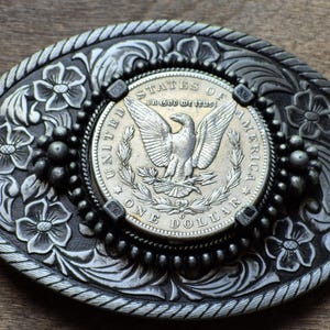 May include: Silver-coloured oval belt buckle with floral and rope detailing. The centre features a silver dollar coin with an eagle and the words "United States of America" and "One Dollar".