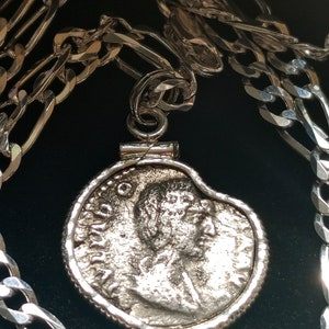 May include: A silver chain necklace with a pendant featuring a silver coin with a profile of a person's face. The coin has a raised rim and intricate details.