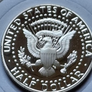 K&ouml;nnte beinhalten: Nahaufnahme einer silbernen Kennedy-Halbdollar-M&uuml;nze. Die M&uuml;nze tr&auml;gt die Aufschrift &bdquo;UNITED STATES OF AMERICA&ldquo; und &bdquo;HALF DOLLAR&ldquo; am Rand. In der Mitte der M&uuml;nze ist ein Adler mit ausgebreiteten Fl&uuml;geln und einem Schild abgebildet.