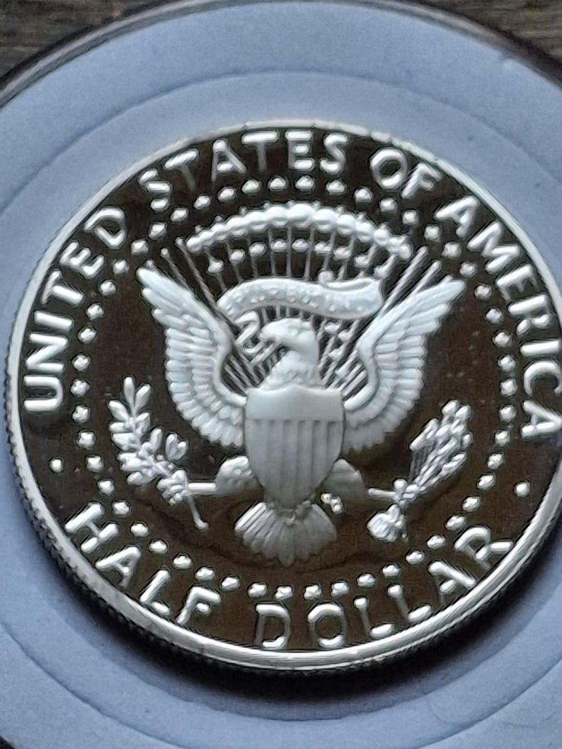 K&ouml;nnte beinhalten: Nahaufnahme einer silbernen Kennedy-Halbdollar-M&uuml;nze. Die M&uuml;nze tr&auml;gt die Aufschrift &bdquo;UNITED STATES OF AMERICA&ldquo; und &bdquo;HALF DOLLAR&ldquo; am Rand. In der Mitte der M&uuml;nze ist ein Adler mit ausgebreiteten Fl&uuml;geln und einem Schild abgebildet.