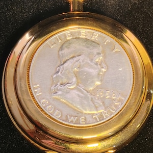 May include: A gold-toned pocket watch with a silver-colored face featuring a profile of a person and the text "LIBERTY", "IN GOD WE TRUST", and "1958". The watch has a circular design and a loop at the top.