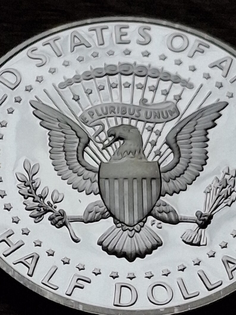 K&ouml;nnte beinhalten: Nahaufnahme einer silbernen Kennedy-Halbdollar-M&uuml;nze. Die M&uuml;nze zeigt die Worte "UNITED STATES OF AMERICA" und "HALF DOLLAR" am Rand. In der Mitte der M&uuml;nze ist ein Adler mit ausgebreiteten Fl&uuml;geln zu sehen, der einen Olivenzweig und Pfeile h&auml;lt.