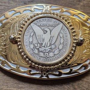 Peut inclure: Boucle de ceinture ovale en métal doré avec une pièce de monnaie argentée au centre. La pièce représente un aigle et les mots "UNITED STATES OF AMERICA" et "ONE DOLLAR". La boucle présente des détails floraux et perlés.