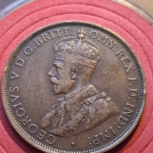 May include: A bronze coin featuring a profile of a man wearing a crown, with text around the edge. The coin is set against a red background. The text includes "GEORGIVS V D.G. BRITT: OMN: REX F.D. IND: IMP:"
