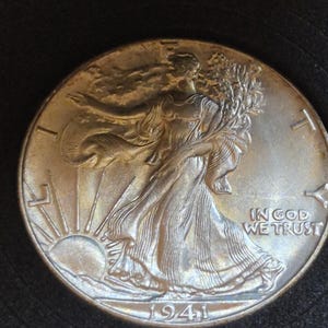 May include: A silver-toned Walking Liberty half dollar coin dated 1941. The obverse features a figure draped in a flag walking toward the left, with the word LIBERTY arched above, the phrase IN GOD WE TRUST to the right, and the sun rising below.