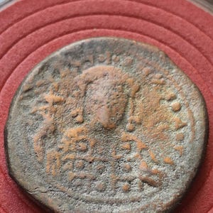 May include: A worn, bronze coin with detailed relief. The coin displays a central bust and surrounding inscriptions. The coin's surface shows signs of age and wear, with a reddish-brown hue. The coin is set against a red background.