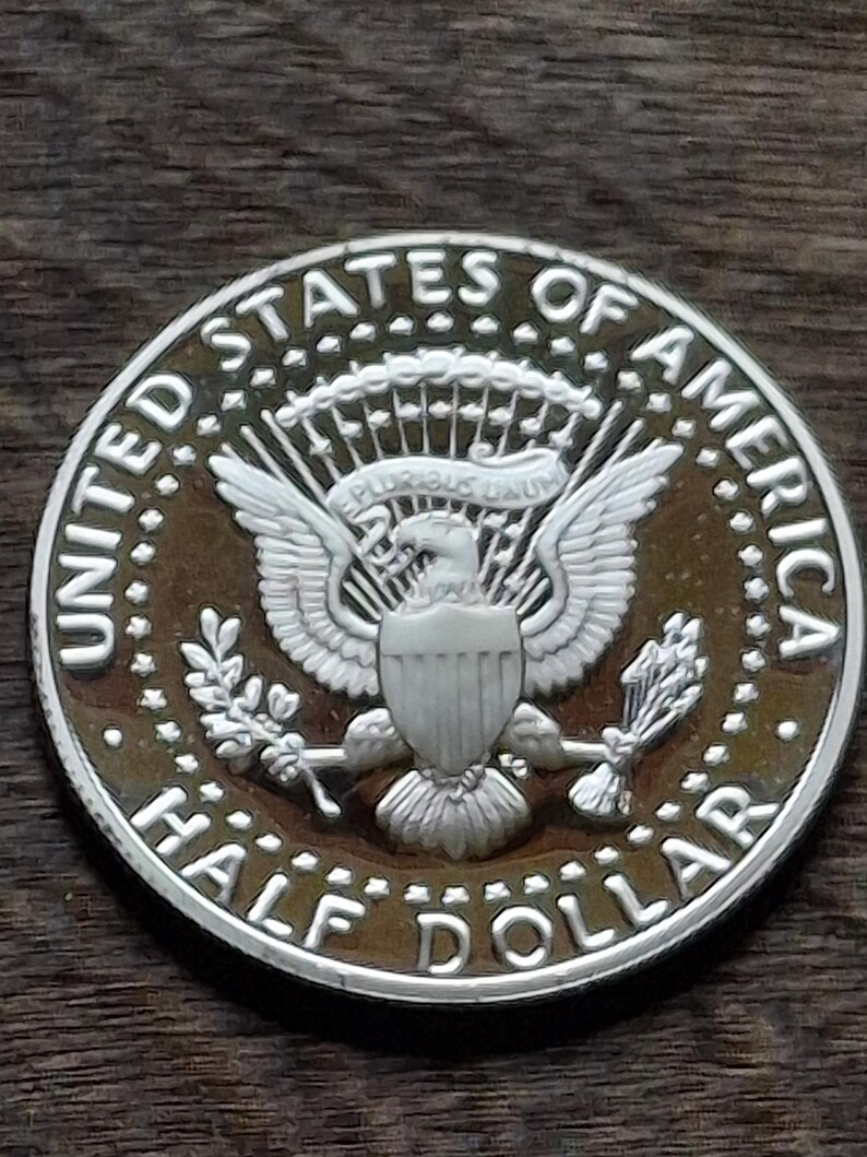 K&ouml;nnte beinhalten: Eine silberne Half-Dollar-M&uuml;nze mit detailliertem Design. Die M&uuml;nze tr&auml;gt die Aufschrift "UNITED STATES OF AMERICA" und "HALF DOLLAR". In der Mitte ist ein Adler mit ausgebreiteten Fl&uuml;geln zu sehen, der Pfeile und einen Olivenzweig h&auml;lt.