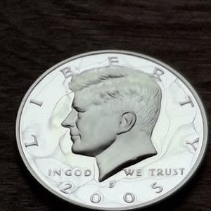 K&ouml;nnte beinhalten: Eine silberne Kennedy-Halbdollar-M&uuml;nze von 2005. Die M&uuml;nze zeigt ein Profil von John F. Kennedy und die Inschriften "LIBERTY", "IN GOD WE TRUST" und "UNITED STATES OF AMERICA". Die M&uuml;nze liegt auf einer dunkelbraunen Oberfl&auml;che.