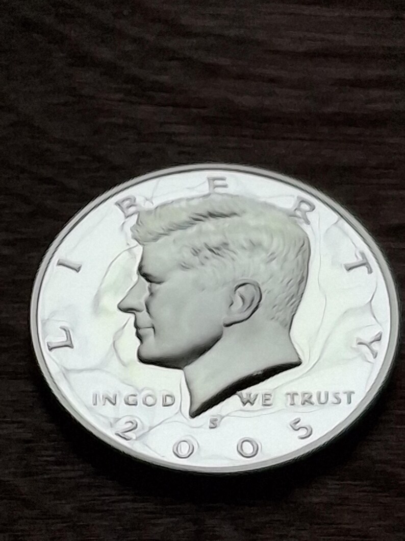 K&ouml;nnte beinhalten: Eine silberne Kennedy-Halbdollar-M&uuml;nze von 2005. Die M&uuml;nze zeigt ein Profil von John F. Kennedy und die Inschriften "LIBERTY", "IN GOD WE TRUST" und "UNITED STATES OF AMERICA". Die M&uuml;nze liegt auf einer dunkelbraunen Oberfl&auml;che.
