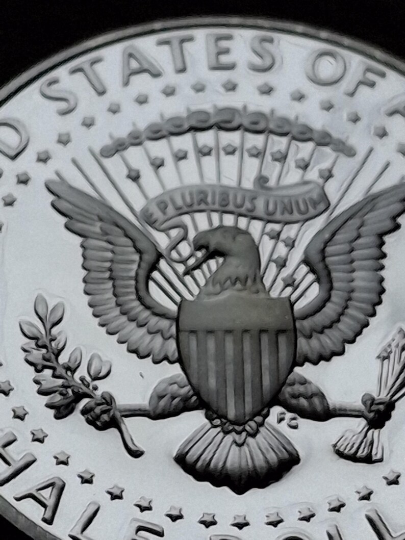 K&ouml;nnte beinhalten: Nahaufnahme einer Silberm&uuml;nze mit dem Gro&szlig;en Siegel der Vereinigten Staaten. Das Design umfasst einen Adler mit ausgebreiteten Fl&uuml;geln, einen Schild und die Inschrift "E PLURIBUS UNUM". Die M&uuml;nze tr&auml;gt auch die Worte "UNITED STATES OF" und "HALF DOLLAR".