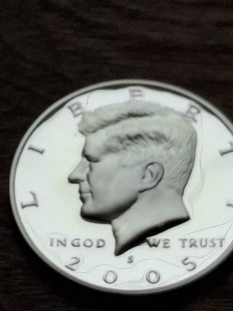 K&ouml;nnte beinhalten: Nahaufnahme einer silbernen Kennedy-Halbdollar-M&uuml;nze von 2005. Die M&uuml;nze zeigt ein Profil von John F. Kennedy und die Worte "IN GOD WE TRUST". Die M&uuml;nze befindet sich auf einem dunklen Hintergrund.