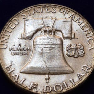 May include: A silver-coloured United States half dollar coin featuring the Liberty Bell in the centre. The text UNITED STATES OF AMERICA and HALF DOLLAR surrounds the bell. The phrase E PLURIBUS UNUM and a small eagle are also visible on the coin.
