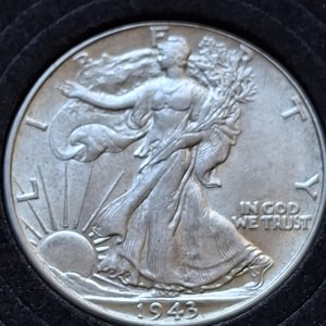 May include: A silver 1943 Walking Liberty half dollar coin. The coin depicts Lady Liberty walking towards the sun with an olive branch. The inscription "IN GOD WE TRUST" is visible, along with the year.