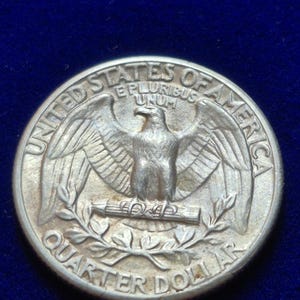 Puede incluir: Una moneda de un cuarto de dólar de los Estados Unidos de color plateado con un águila de alas extendidas posada sobre un haz de flechas. El texto UNITED STATES OF AMERICA, QUARTER DOLLAR y E PLURIBUS UNUM está grabado en la moneda circular.