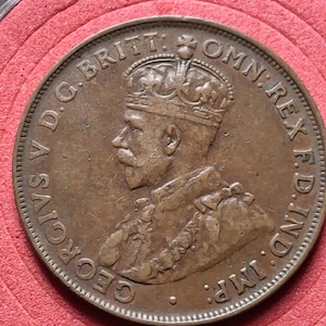 Könnte beinhalten: Eine Bronzemünze mit dem Profil eines Mannes mit Krone und Text am Rand. Die Münze ist vor einem roten Hintergrund platziert. Der Text enthält "GEORGIVS V D.G. BRITT: OMN: REX F.D. IND: IMP:"