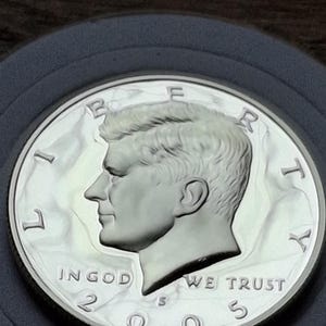 K&ouml;nnte beinhalten: Eine silberne Kennedy-Halbdollar-M&uuml;nze von 2005. Die M&uuml;nze zeigt ein Profil von John F. Kennedy und die Worte "IN GOD WE TRUST". Die M&uuml;nze befindet sich in einer Schutzh&uuml;lle.