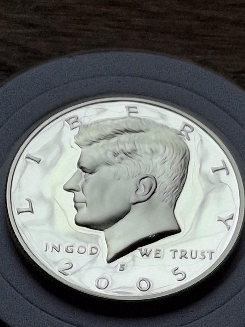 K&ouml;nnte beinhalten: Eine silberne Kennedy-Halbdollar-M&uuml;nze von 2005. Die M&uuml;nze zeigt ein Profil von John F. Kennedy und die Worte "IN GOD WE TRUST". Die M&uuml;nze befindet sich in einer Schutzh&uuml;lle.