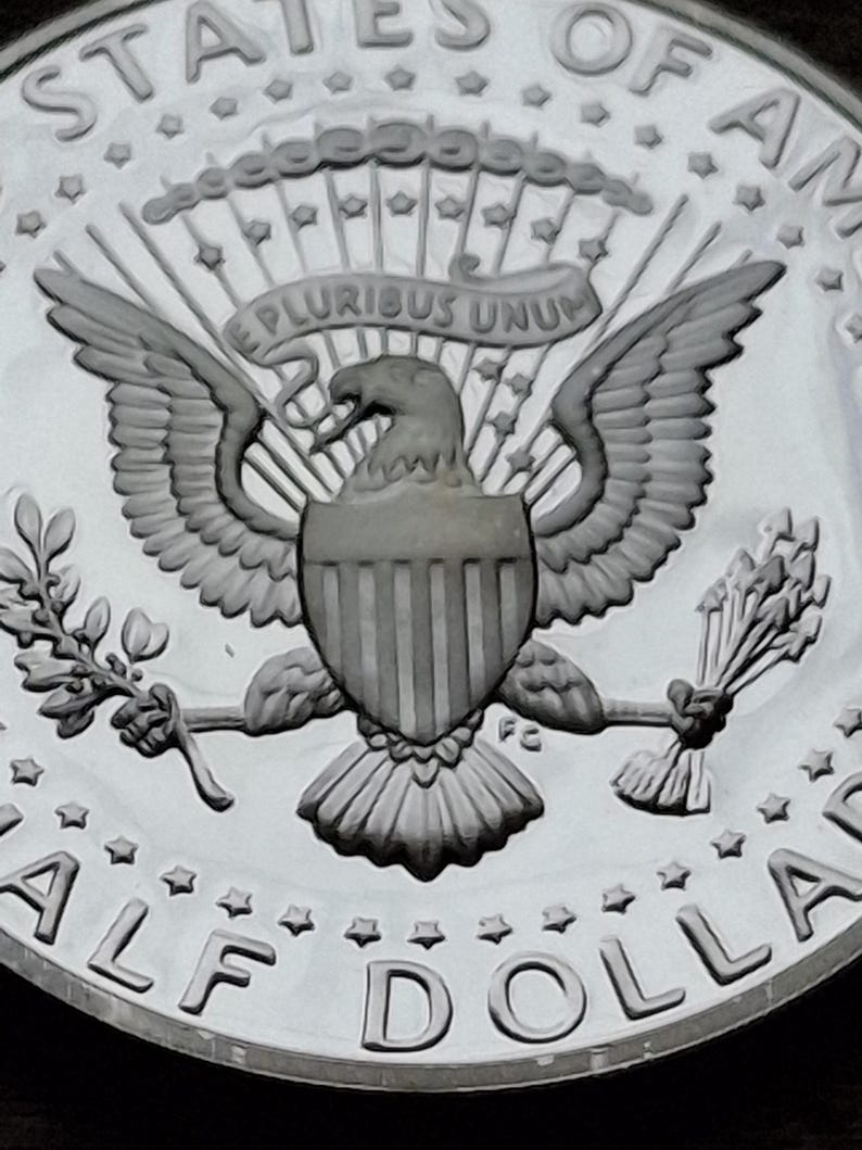 K&ouml;nnte beinhalten: Nahaufnahme einer silbernen Half-Dollar-M&uuml;nze. Die M&uuml;nze zeigt eine detaillierte Darstellung eines Adlers mit ausgebreiteten Fl&uuml;geln, der einen Olivenzweig und Pfeile h&auml;lt. Die Worte "E PLURIBUS UNUM" sind auf einem Banner &uuml;ber dem Adler eingraviert. Der Rand der M&uuml;nze lautet "HALF DOLLAR".