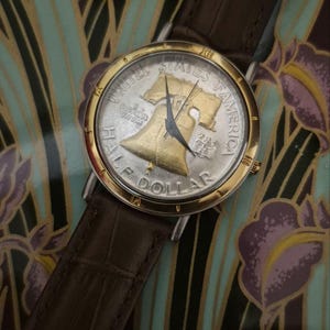 May include: A wristwatch with a silver and gold face featuring a half-dollar coin design. The watch has a brown leather band and a gold-colored bezel. The coin design includes the words "United States of America" and "Half Dollar."