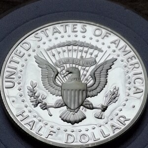 K&ouml;nnte beinhalten: Eine silberne Half-Dollar-M&uuml;nze mit den Worten "UNITED STATES OF AMERICA" und "HALF DOLLAR" am Rand. Die M&uuml;nze zeigt einen Adler mit ausgebreiteten Fl&uuml;geln, der einen Olivenzweig und Pfeile h&auml;lt.