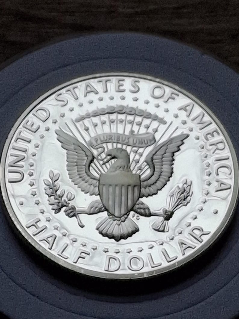 K&ouml;nnte beinhalten: Eine silberne Half-Dollar-M&uuml;nze mit den Worten "UNITED STATES OF AMERICA" und "HALF DOLLAR" am Rand. Die M&uuml;nze zeigt einen Adler mit ausgebreiteten Fl&uuml;geln, der einen Olivenzweig und Pfeile h&auml;lt.