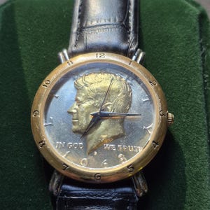 May include: A gold-toned wristwatch featuring a silver coin with a profile of John F. Kennedy. The watch has a black leather band and a gold bezel. The coin face displays the words "IN GOD WE TRUST".
