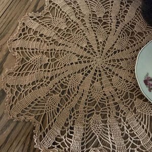 May include: A round, tan-colored, crocheted doily with a detailed openwork design. The doily has a scalloped edge and is placed on a wooden surface. A portion of a teacup and a blue glass object are visible.