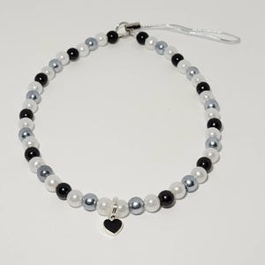 May include: A black and white beaded phone charm with a silver heart pendant. The charm is made with black, white, and silver beads.