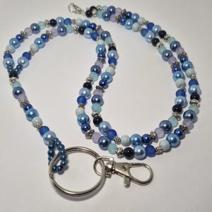 Peut inclure: Un cordon porte-badge composé de perles bleues, bleu marine et violet clair aux finitions mates, brillantes et nacrées. Les perles sont agrémentées de petites entretoises argentées et reliées à un anneau fendu et un mousqueton en métal argenté.