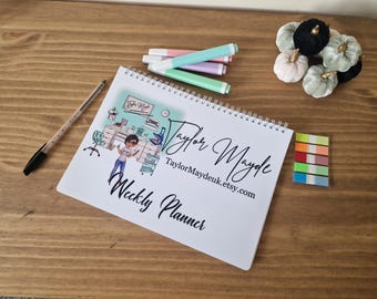May include: A white weekly planner with a spiral binding. The cover features a cartoon illustration of a woman in a lab coat and the text "Taylor Mayde Weekly Planner" and "TaylorMaydeuk.etsy.com".