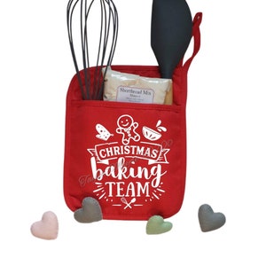 Könnte beinhalten: Ein roter Ofenhandschuh mit einem weißen Design, auf dem "Christmas Baking Team" mit einem Lebkuchenmann, einem Schneebesen und einer Schüssel steht. Der Ofenhandschuh hat zwei Taschen, eine mit einem Schneebesen und die andere mit einem Spatel. In der Tasche mit dem Spatel befindet sich eine Tüte Shortbread-Mix.