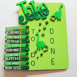 May include: A green wooden board with the name "Jake" written in green letters. The board has a dinosaur cutout and a checklist with the words "Brush Teeth", "Get Dressed", "Make Bed", "Tidy Toys Away", "Tidy Room", "Remember Manners", "Tidy Dinner Things", "Bath", "Kind Hands", and "Brush Teeth".