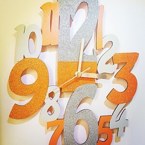 May include: A decorative wall clock with large, glittery orange, silver, and white numbers. The clock hands are gold, and the numbers are arranged in a circular pattern. The clock face is a mix of orange and silver glitter.