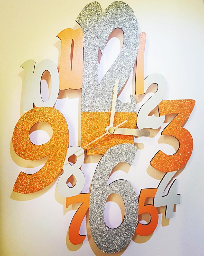 May include: A decorative wall clock with large, glittery orange, silver, and white numbers. The clock hands are gold, and the numbers are arranged in a circular pattern. The clock face is a mix of orange and silver glitter.