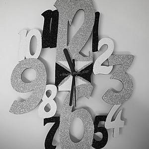 May include: A decorative wall clock with large, glittery numbers in silver, black, and white. The clock face is a geometric design in black and white. The hands are black. The clock is mounted on a white wall.