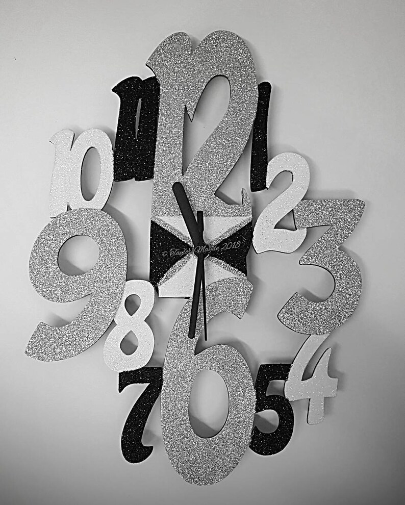 May include: A decorative wall clock with large, glittery numbers in silver, black, and white. The clock face is a geometric design in black and white. The hands are black. The clock is mounted on a white wall.