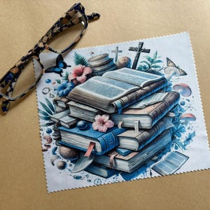 May include: A white microfiber cleaning cloth with a colorful illustration of a stack of books, butterflies, flowers, and other decorative elements. The cloth is designed for cleaning glasses.