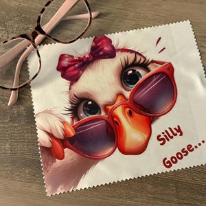 May include: A white microfiber cleaning cloth featuring a cartoon goose wearing red sunglasses and a red bow. The text "Silly Goose..." is printed on the cloth. A pair of eyeglasses with a brown and pink frame rests on the wooden surface.