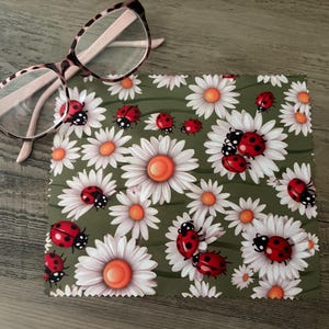May include: A pair of eyeglasses with a pink frame rests on a green cloth. The cloth is patterned with white daisies and red ladybugs. The eyeglasses have a tortoiseshell pattern on the frame.