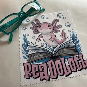 May include: A white microfiber cleaning cloth with a pink axolotl sitting on a book. The axolotl is surrounded by bubbles and seaweed. The text "Read Axolotl" is printed in pink on the cloth.