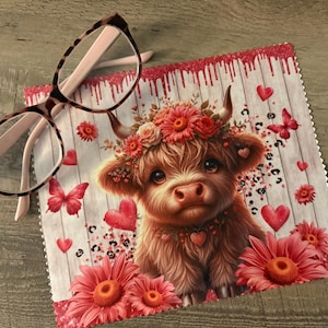 May include: A microfiber cleaning cloth featuring a Highland cow illustration adorned with a floral crown, hearts, and butterflies. The cloth is decorated with pink and red accents, and a pair of eyeglasses rests on top.