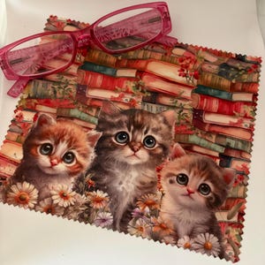 May include: Pink eyeglasses rest on a patterned cloth featuring three adorable kittens surrounded by flowers, set against a backdrop of stacked books. The glasses have a rectangular frame.