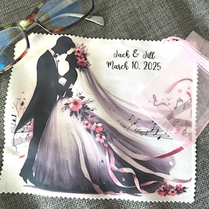 Op de afbeelding: Een witte microvezel schoonmaakdoek met een aquarel-illustratie van een bruid en bruidegom in silhouet. De doek heeft een roze rand en de tekst "Jack & Jill March 10, 2025" erop gedrukt.