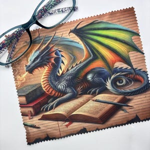 May include: A square microfibre cleaning cloth with pinked edges featuring a colourful illustration of a dragon with green and orange wings resting on open books. A pair of glasses with a patterned frame rests on the top left corner of the cloth.