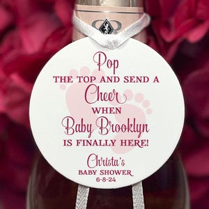May include: White circle bottle topper with pink footprints and the text "Pop the top and send a cheer when Baby Brooklyn is finally here! Christa's Baby Shower 6-8-24"