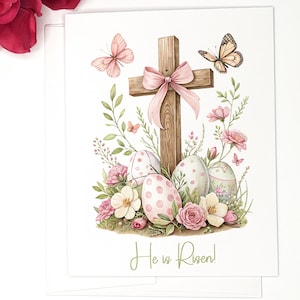 May include: A watercolor illustration of a wooden cross with a pink ribbon, surrounded by pink and white flowers, green leaves, and pastel Easter eggs. The text "He is Risen!" is written below the cross.