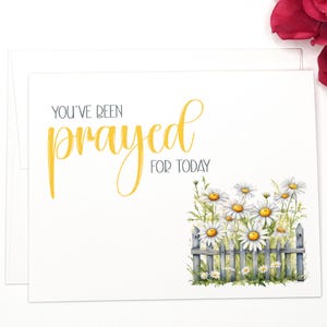 May include: A white card with the text "You've been prayed for today" in yellow lettering. The card features a watercolor illustration of a white picket fence with white daisies.