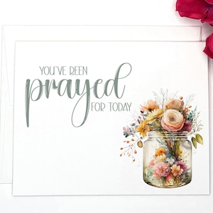 May include: A white greeting card with the text "YOU'VE BEEN prayed FOR TODAY" in green script. The card features a watercolor illustration of a mason jar filled with colorful flowers, including roses and daisies. The card is blank inside.