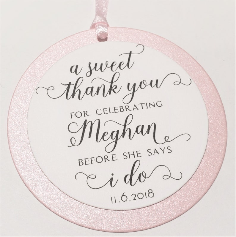 Pode incluir: Uma etiqueta de favor redonda, rosa e com glitter com um c&iacute;rculo branco no centro. A etiqueta diz "a sweet thank you for celebrating Meghan before she says i do 11.6.2018".