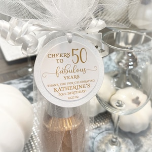 May include: White round favor tag with gold lettering. It says "Cheers to 50 fabulous years. Thank you for celebrating Katherine's 50th Birthday! 10.22.22".